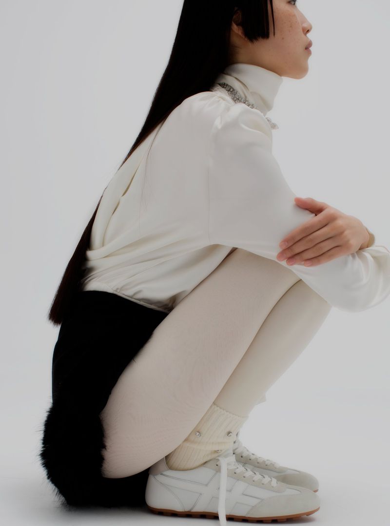 A slender adult female poses in a crouched position with long, dark hair in a white long-sleeved shirt, cream leggings, and vintage sneakers.