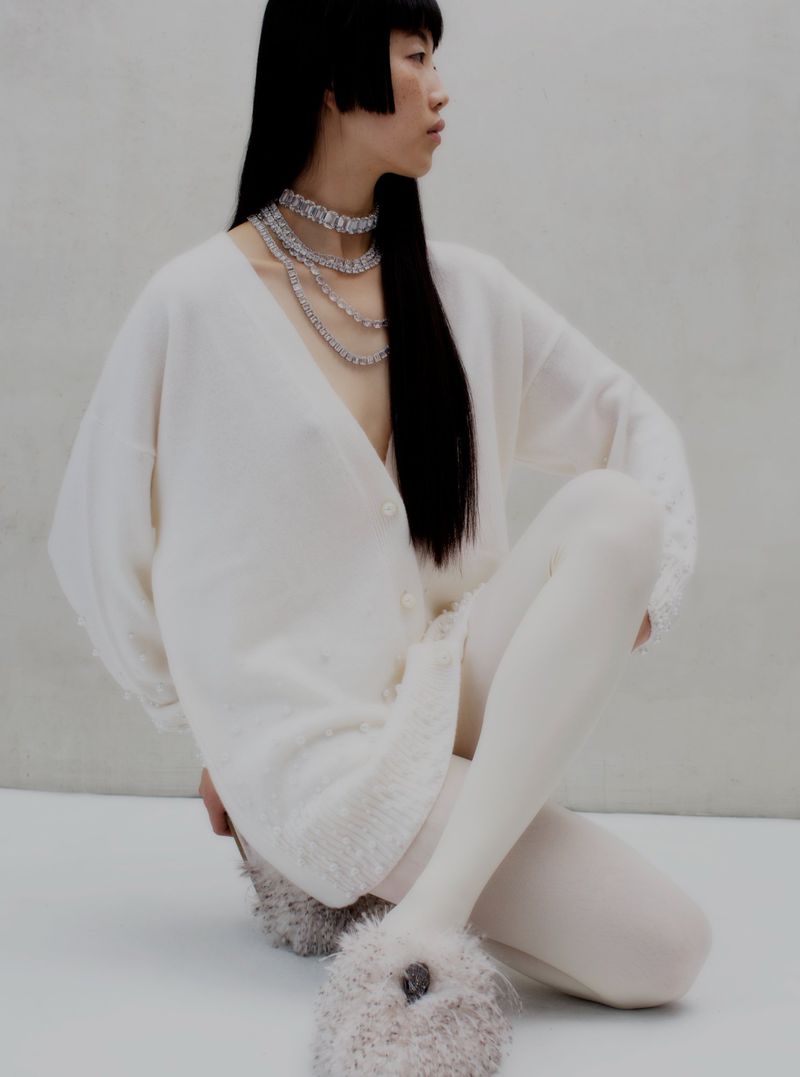 Asian woman in fashion shot wearing white cardigan sweater, white tights, layered necklace, and fluffy slippers against gray backdrop.