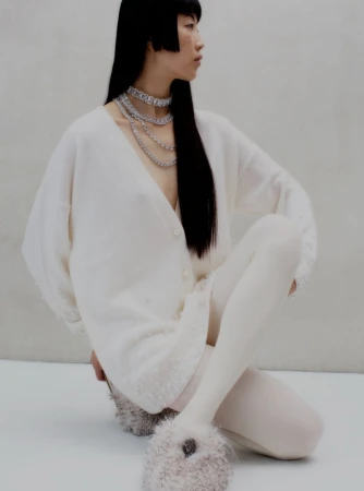 Asian woman in fashion shot wearing white cardigan sweater, white tights, layered necklace, and fluffy slippers against gray backdrop.