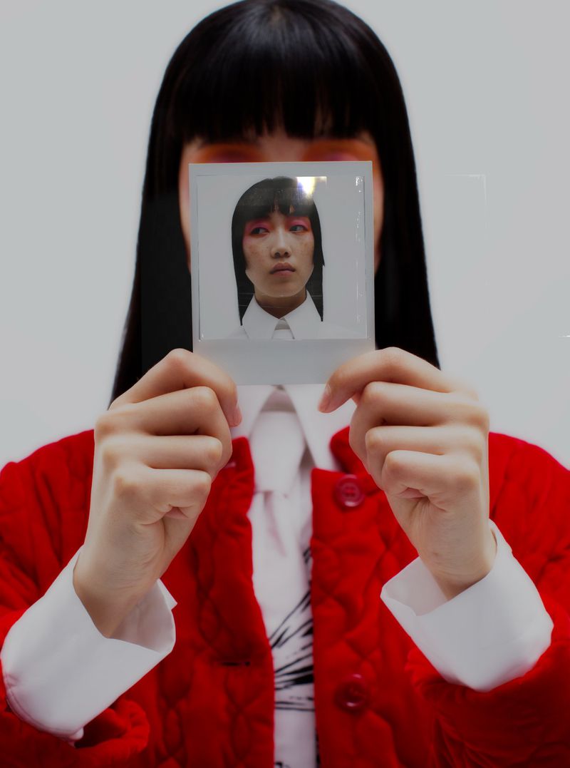 Woman holds instant photo of another person's face, wearing a red quilted jacket with white background, in a medium studio shot.