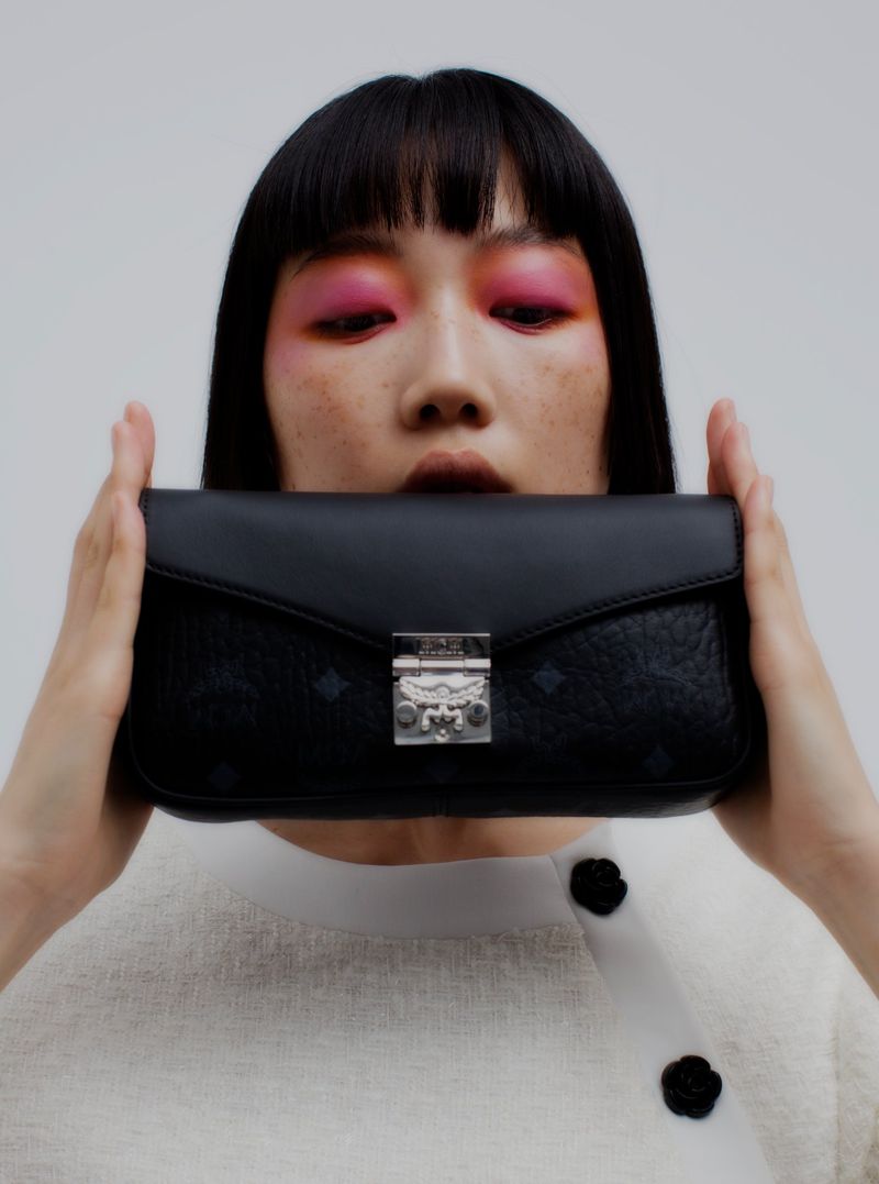 A woman with vibrant pink eyeshadow holds a black MCM handbag in front of her face against a plain, bright background.