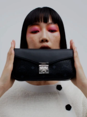 A woman with vibrant pink eyeshadow holds a black MCM handbag in front of her face against a plain, bright background.