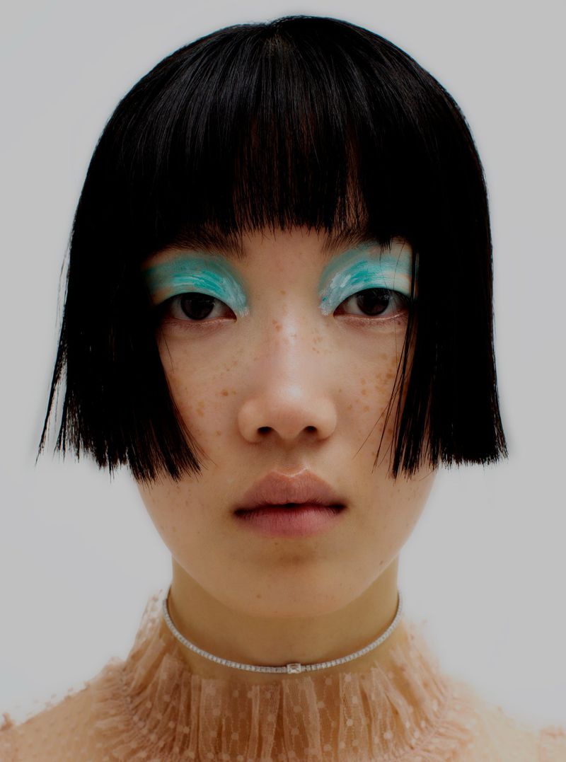 Headshot of a young Asian woman with short black hair and bright turquoise eyeshadow, wearing a diamond choker, against a white background.