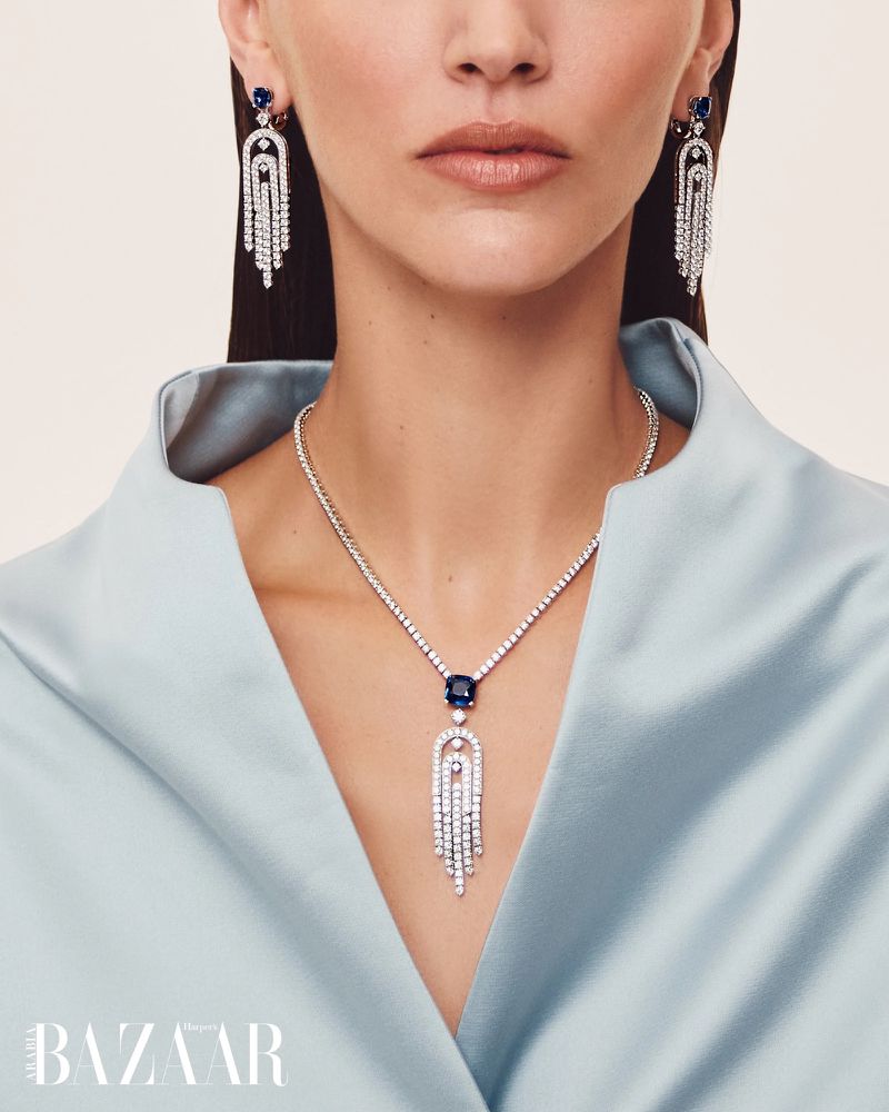 Close-up of a woman wearing a diamond and sapphire necklace and matching earrings, against a soft light blue background in a studio setting.