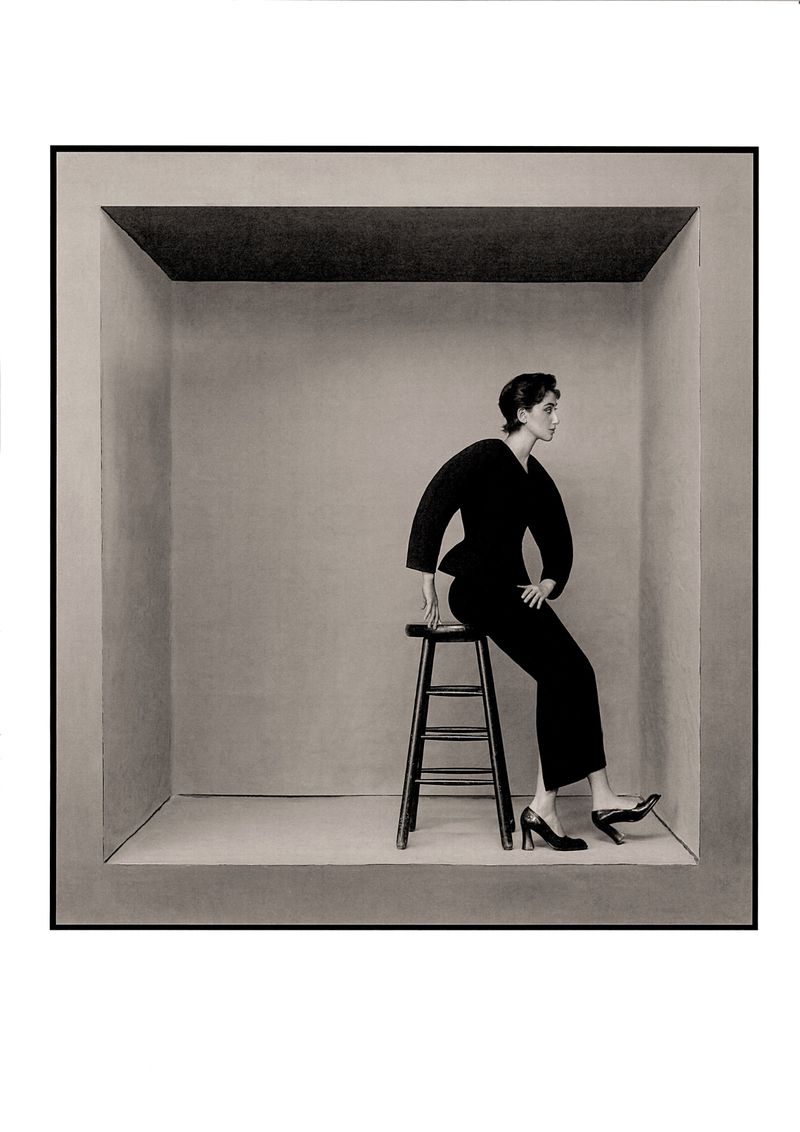 Monochrome portrait of an adult female in a dark pantsuit, sitting on a stool inside a geometric inset against a light gray background.
