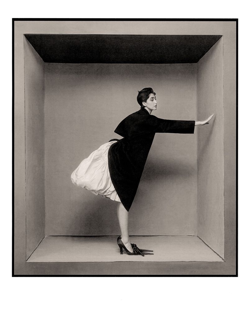 A woman in a black coat and white dress is posing in a gray box with her hand on the wall. High fashion photography with monochrome palette.