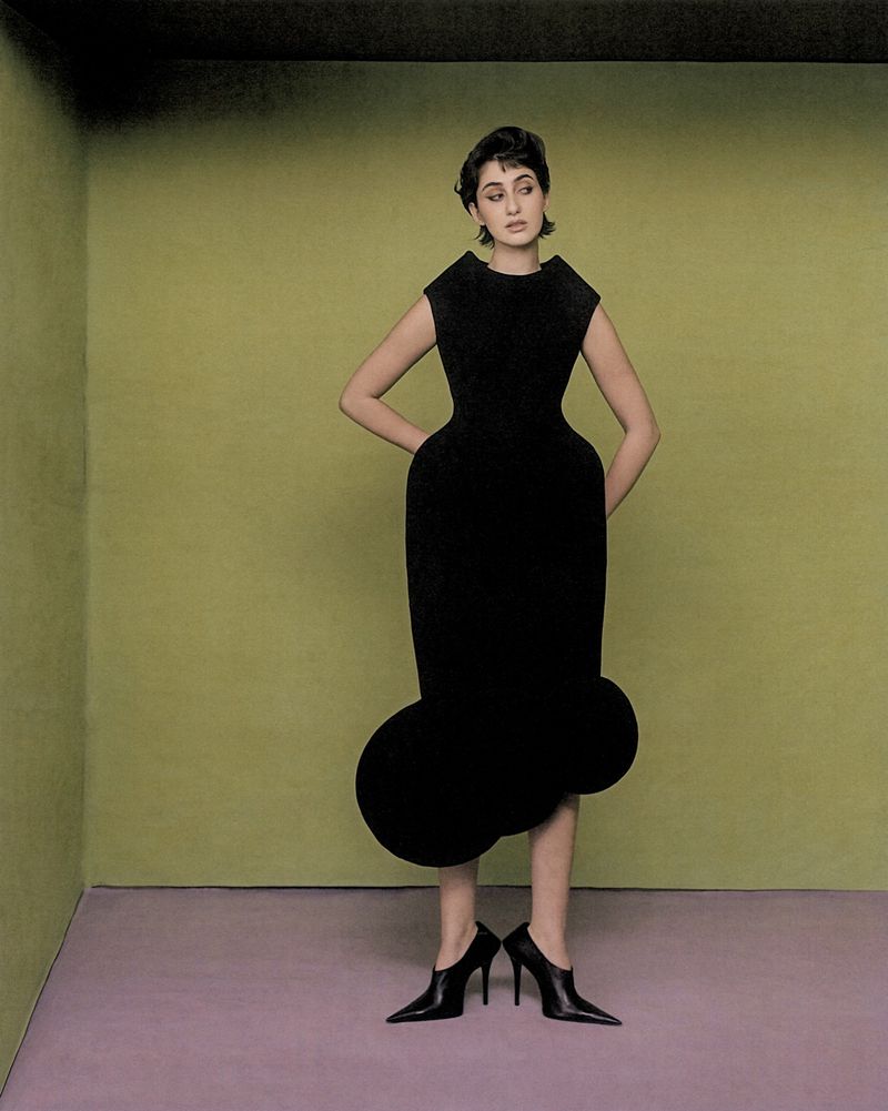 A woman in a black dress and high heels stands in a minimalist room with a green wall and lilac floor, fashion portrait.