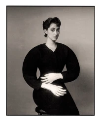Rama Duwaji in a structured black balloon-sleeve dress with white-painted hands resting at her waist against a grey studio backdrop.