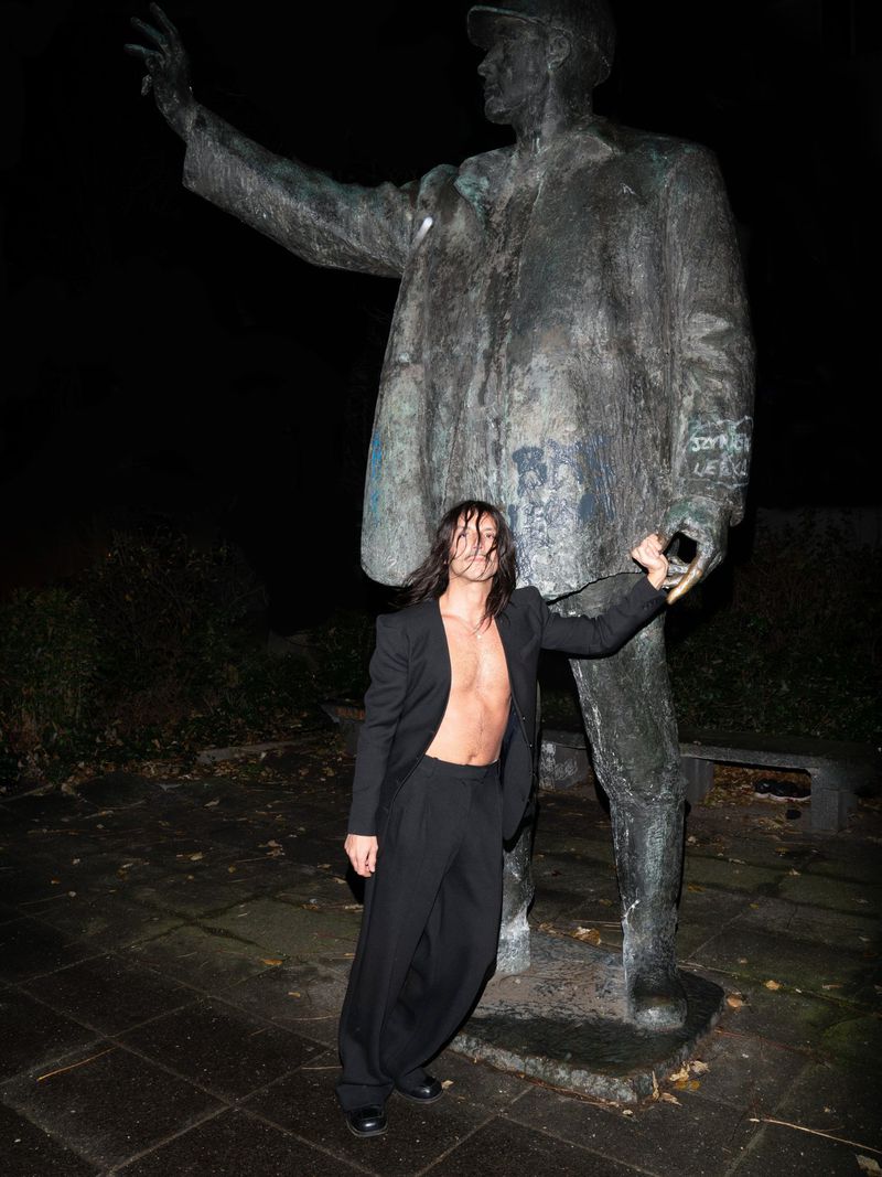 Man with long dark hair wearing an open black suit jacket and trousers standing next to a large weathered metal statue at night.