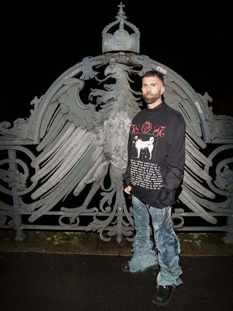 A man stands in front of an ornate, crowned eagle sculpture wearing a black sweater with white graphics and distressed denim jeans.