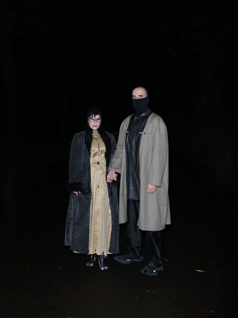 A stylish couple stands together in the dark, the woman in a long coat and the man in a masked face covering and patterned overcoat.