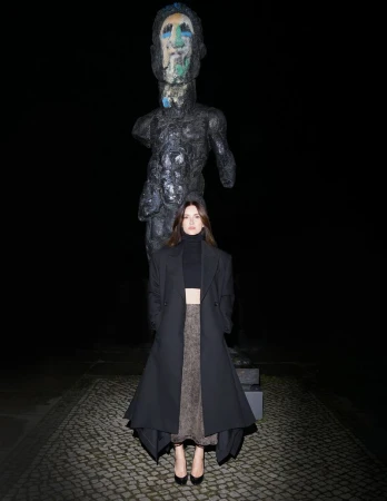 A woman in a black coat standing in front of a sculpture at night, with dark lighting and a cobblestone surface.
