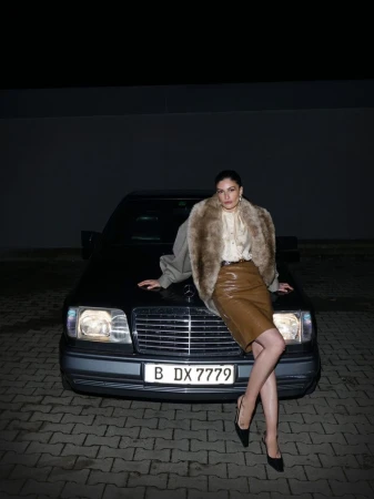 Stylish adult woman in a fur coat poses leaning against a black car on a brick road under dim light, fashion, automotive.