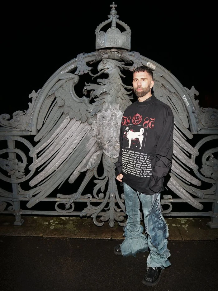 Man in an oversized black graphic long-sleeve shirt and distressed denim jeans standing before an ornate metal eagle gate at night.