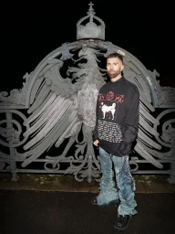 Man in an oversized black graphic long-sleeve shirt and distressed denim jeans standing before an ornate metal eagle gate at night.