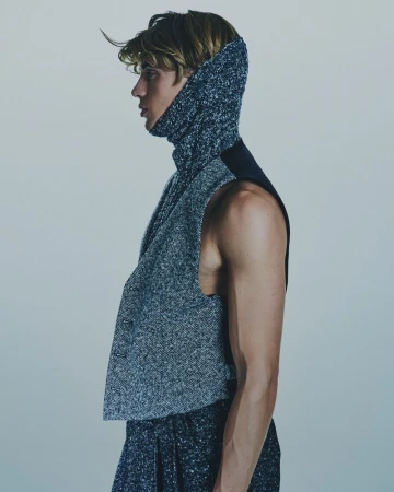 Profile of a blonde adult male fashion model wearing a textured knit hooded scarf and sleeveless top against a grey gradient backdrop.