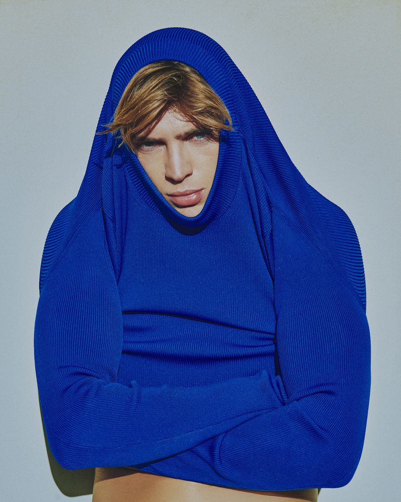 Adult male portrait with fair hair, wearing bright blue knit sweater pulled over his head, posing in a photo studio with a light grey backdrop.