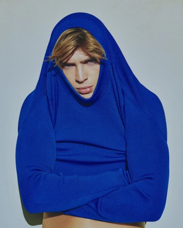 Adult male portrait with fair hair, wearing bright blue knit sweater pulled over his head, posing in a photo studio with a light grey backdrop.