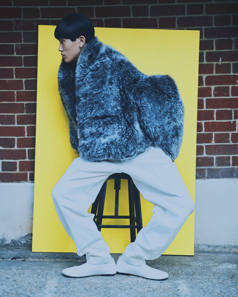 Side view of a person wearing a gray fur coat and white pants, sitting on a stool in front of a yellow backdrop and brick wall.