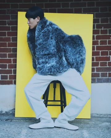 Side view of a person wearing a gray fur coat and white pants, sitting on a stool in front of a yellow backdrop and brick wall.