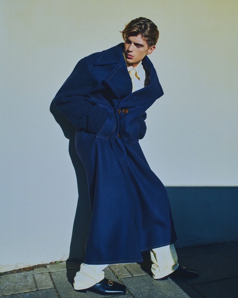 Fashionable young adult in a blue oversized coat with white trousers and black loafers poses next to a white wall outdoors.