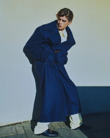 Fashionable young adult in a blue oversized coat with white trousers and black loafers poses next to a white wall outdoors.