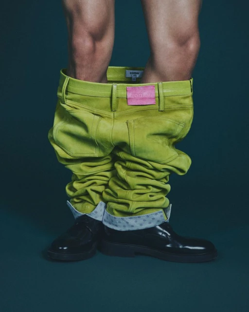 Model displays a pair of bright green pants around ankles, black shiny boots, and a glimpse of skin. Fashion-forward, eye-catching clothing.
