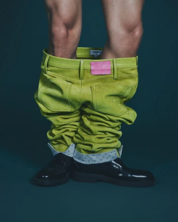 Model displays a pair of bright green pants around ankles, black shiny boots, and a glimpse of skin. Fashion-forward, eye-catching clothing.
