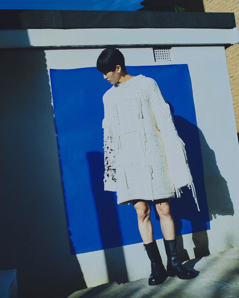 A person in a knitted sweater and boots stands against a blue and white wall on a sunny day, creating shadows. Stylish fashion photo.
