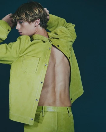 Blonde male model poses in matching unbuttoned chartreuse jacket and pants against a dark teal backdrop in a studio fashion shot.