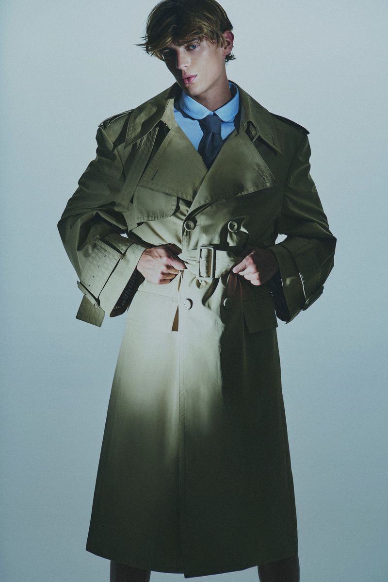 Stylish adult male fashion model poses in a tan trench coat, blue shirt, and tie in a studio setting against a light blue backdrop.
