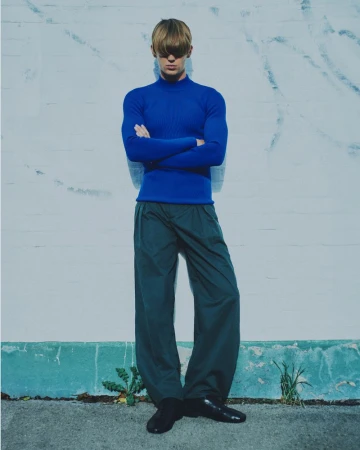 Full-length portrait of an adult male fashion model with crossed arms wearing a blue sweater and green pants against a white brick wall.