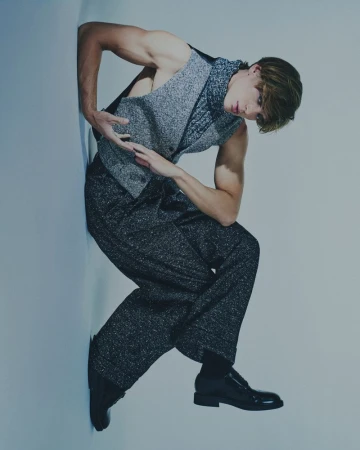 Fashion shot of a young male model with arms raised, wearing a grey vest and pants, leans against a wall in a photo studio.