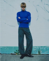 Model with blond bangs in a cobalt blue long-sleeve turtleneck and dark wide-leg trousers standing with arms crossed against a wall.