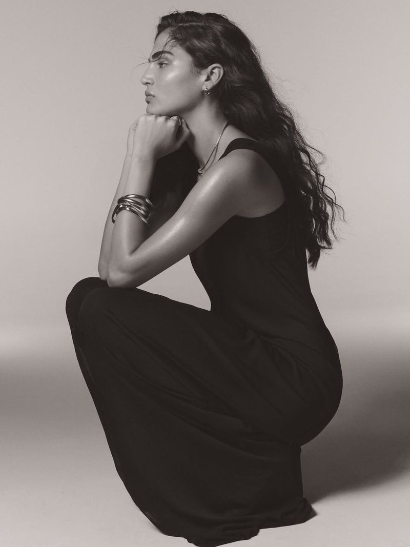 Monochrome portrait of a woman with long, wavy hair wearing a black dress, seated in a studio with hands near her face.