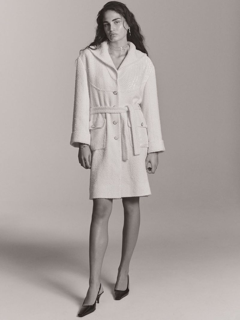 Monochrome fashion portrait of a slender adult woman in a tweed belted coat and slingback heels on a light neutral backdrop.