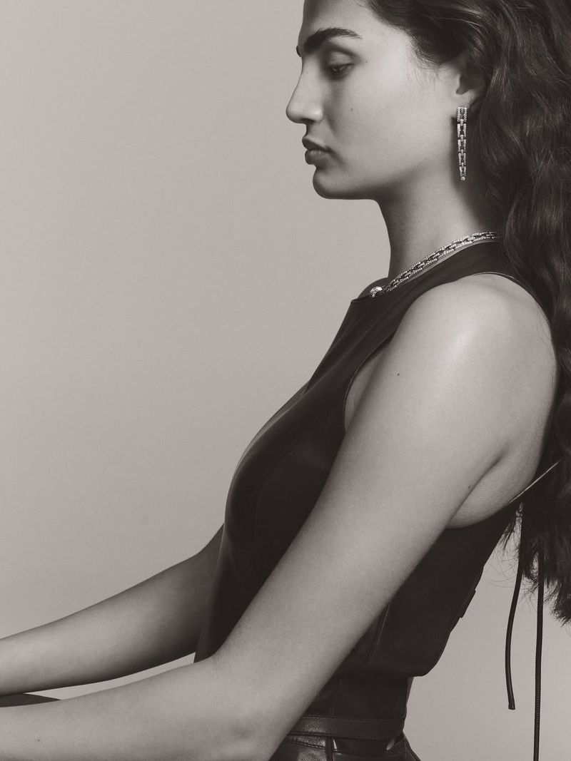 Profile portrait of a woman wearing fine jewellery including drop earrings and a necklace against a plain grey background