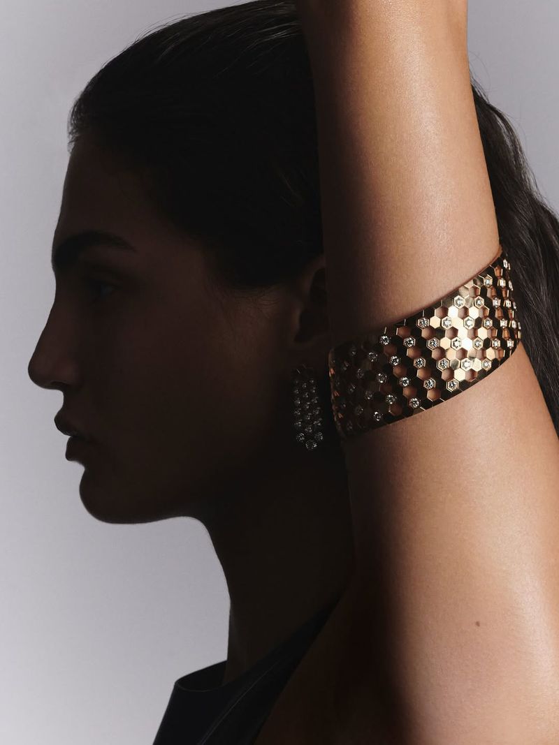 Profile of a woman wearing a honeycomb patterned gold cuff bracelet and matching earrings, against a graduated gray background.