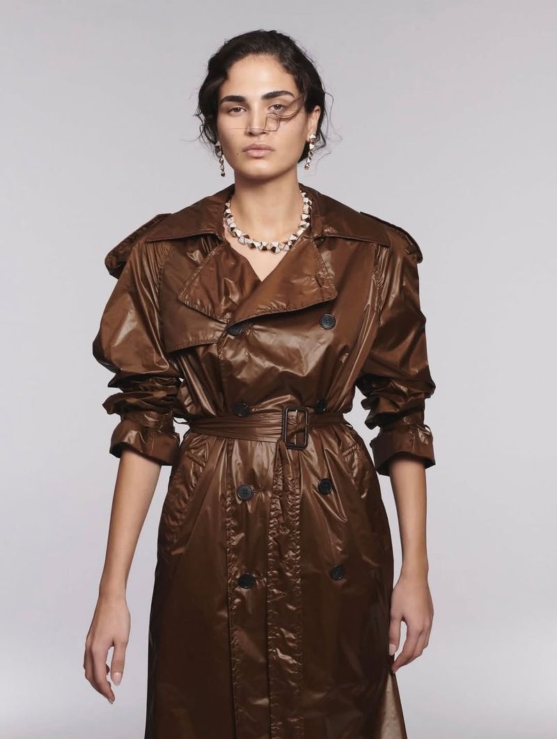 Elegant adult female posing in shiny brown trench coat, accessorized with pearl necklace and earrings, in a studio setting.