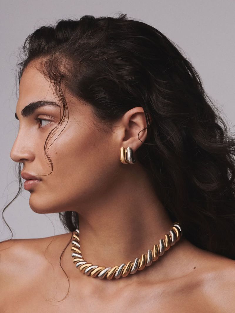 Profile portrait of a woman wearing a two-tone twisted gold and silver necklace and matching earrings against a grey studio backdrop.