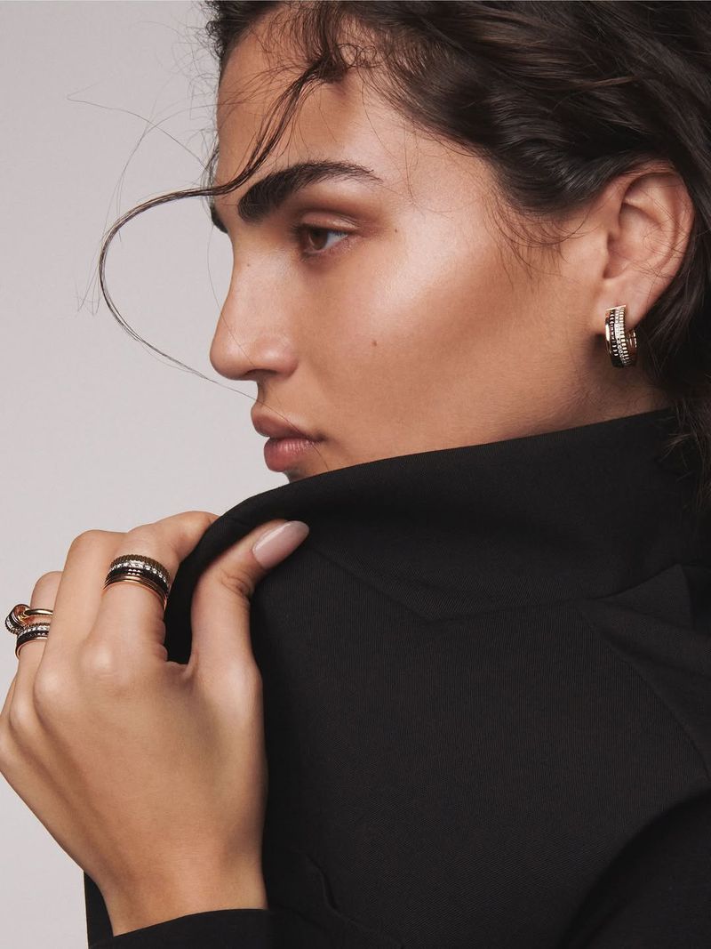 Close-up shot of a brunette adult female wearing gold hoop earrings and multiple gold rings with a neutral expression.