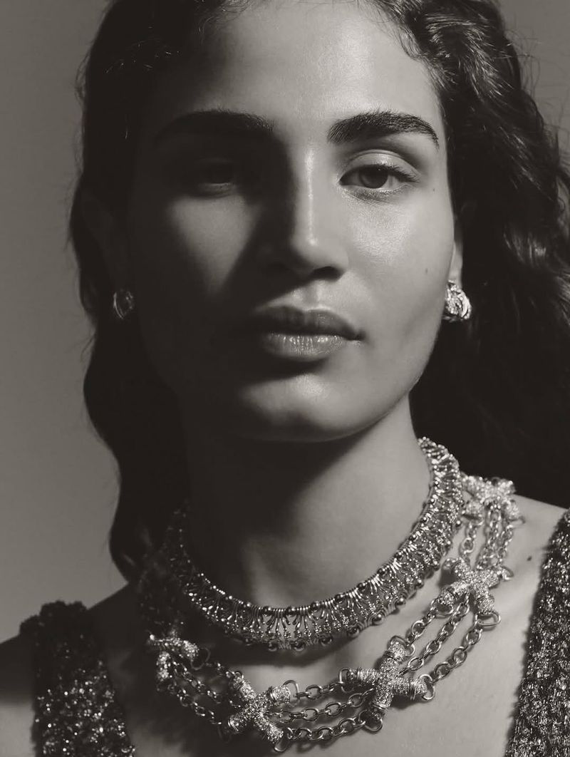 Monochrome portrait of a woman adorned with a layered statement necklace and earrings against a neutral backdrop in soft lighting.