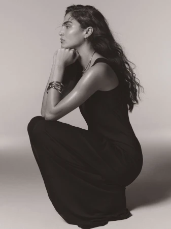 Model crouching in profile wearing a black sleeveless maxi dress and metallic bangles against a seamless studio backdrop.