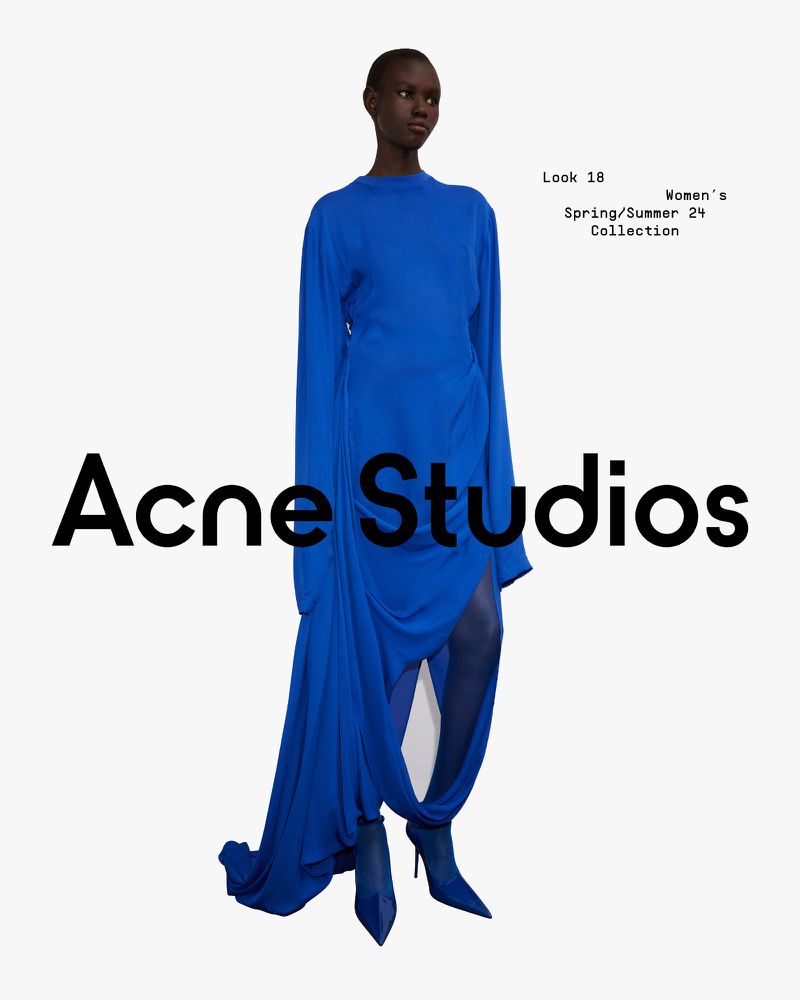 An adult woman poses in a full-length shot wearing a long-sleeved, draped, bright blue dress with matching pointed-toe heeled boots against a white backdrop.