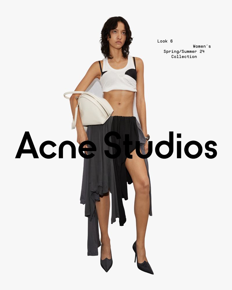 Fashion editorial shot of a woman in a white crop top, black and gray skirt, and heels holding a white bag against a white backdrop.
