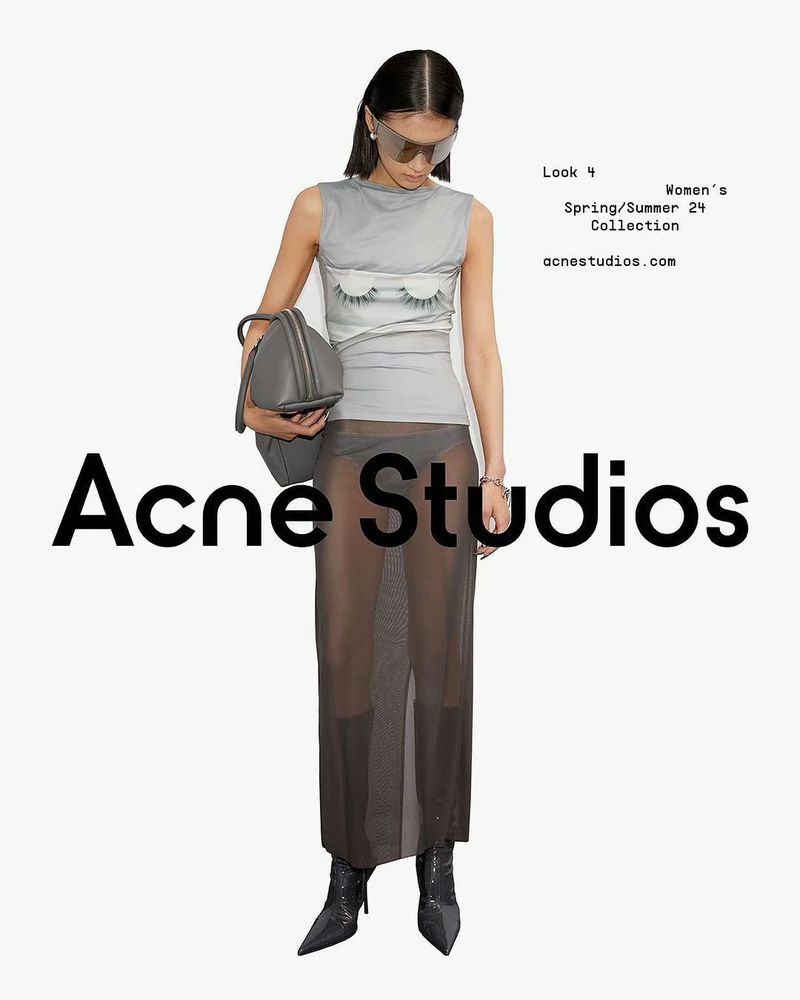 Fashion shot featuring a woman in a sleeveless grey top with an eye graphic, a sheer maxi skirt, sunglasses, boots, and a grey handbag.