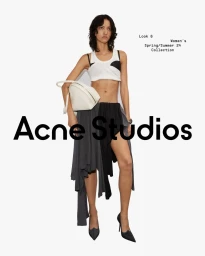 Model wearing white crop top, asymmetrical grey and black skirt, and holding a white handbag against a white background