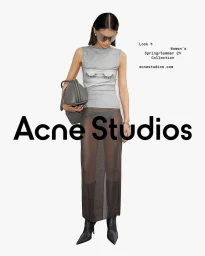 Model standing against a plain white background wearing a sheer skirt, sleeveless top with eyelash graphic, and tinted sunglasses holding a grey handbag