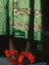 Model Mofeoluwa Onasanya wearing an emerald green embroidered dress and bright red satin bow heels against a dark background.
