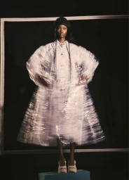 Model Mofeoluwa Onasanya posing in a spinning sheer white dress and black hat against a dark studio background.